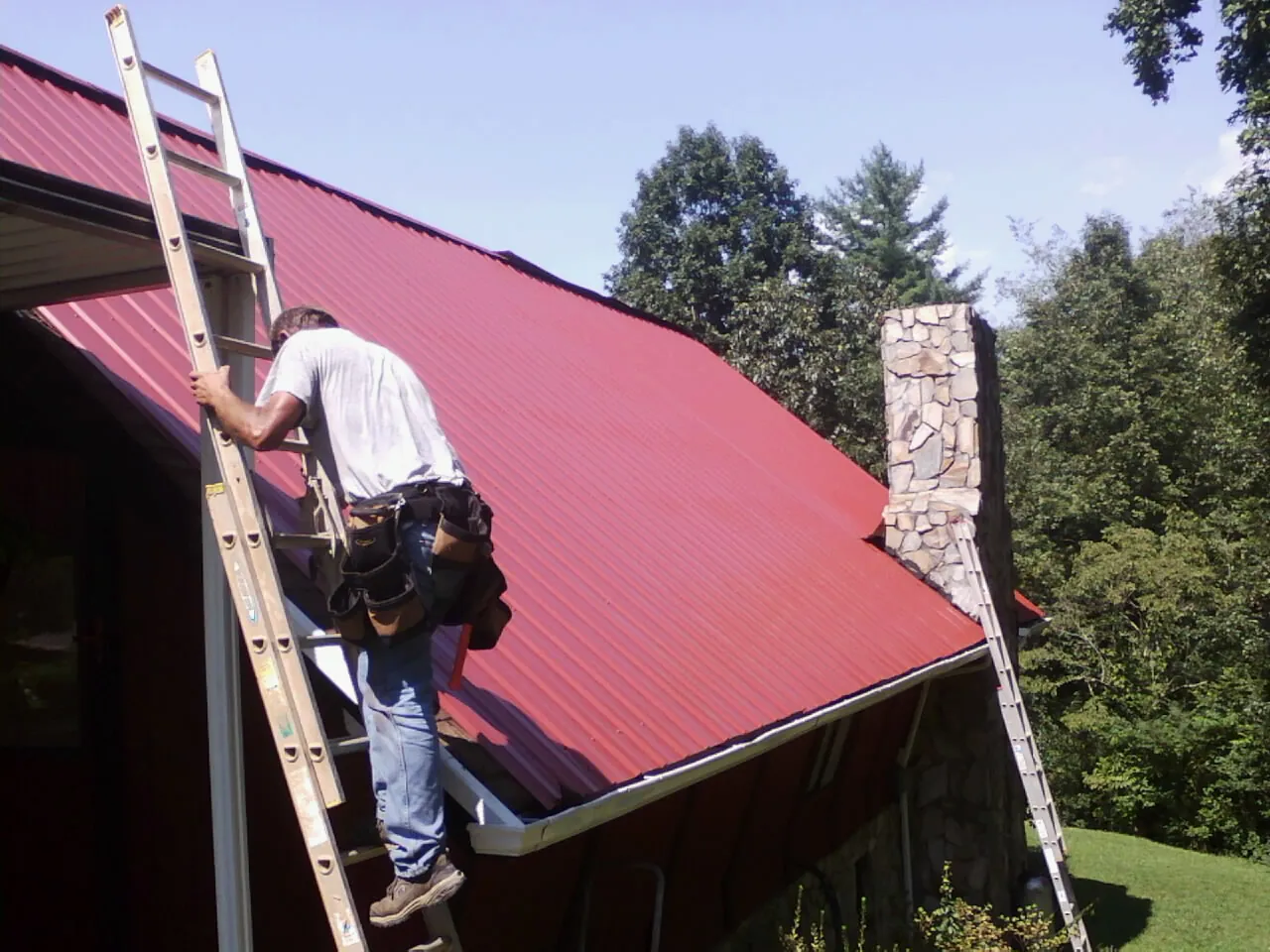 Roofer on a ladder beside a red metal roof and stone chimney — Soffit & Fascia Repair by Roofing Brothers in Clearfield
